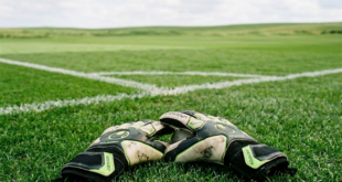 Goalkeeper gloves placed on grass near corner of a soccer field with empty stadium seats in background