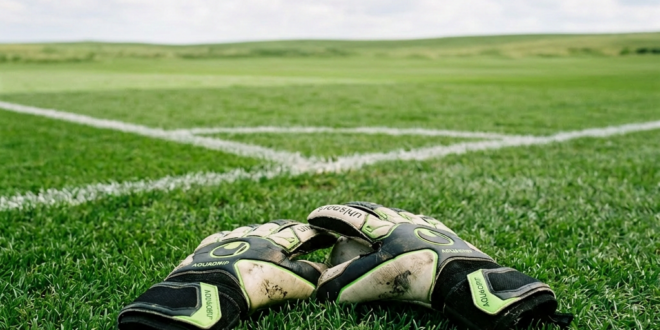 Goalkeeper gloves placed on grass near corner of a soccer field with empty stadium seats in background
